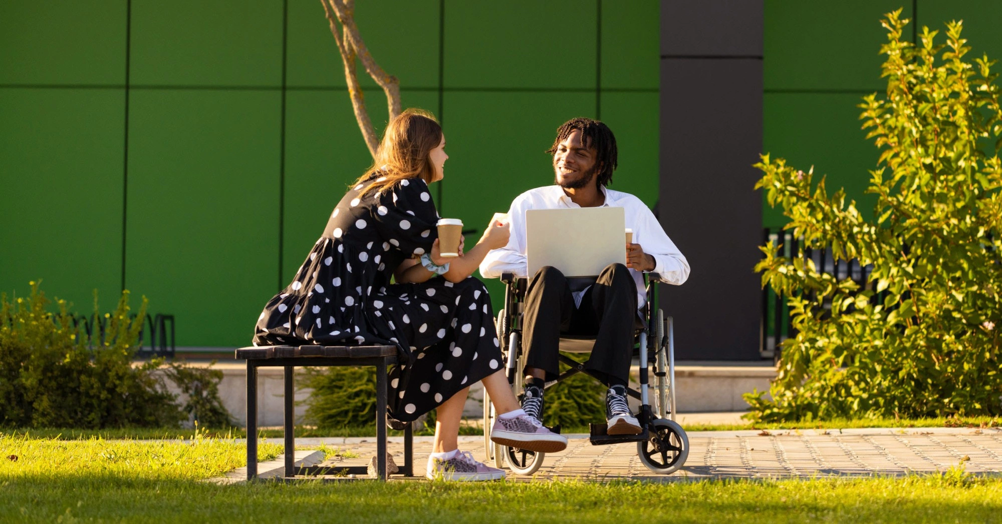 Photo of a person in a wheelchair with a laptop talking to someone sitting on a bench